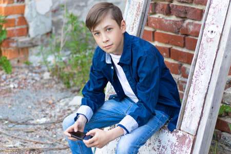 A teenager boy in a blue shirt and jeans with a smartphone in his hands sits on an old door against a brick wall background. Selective focusの写真素材