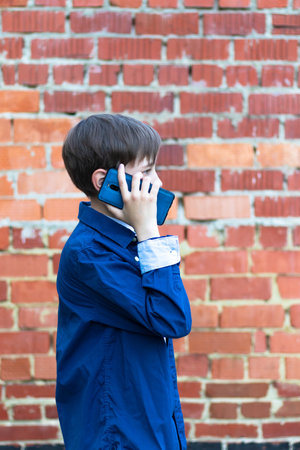 A teenage boy in a blue shirt with a smartphone in his hands communicates with friends on social networks against the background of a brick wall. Selective focusの写真素材
