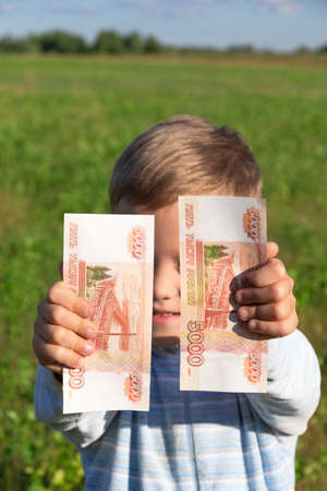 A satisfied child, a preschooler boy, holds paper rubles in his hands outdoors on the background of a field with green grass on a bright sunny summer day. Close-up. Portraitの写真素材