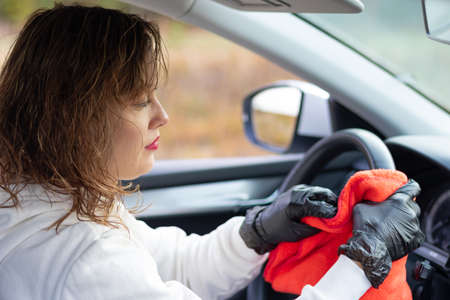 A beautiful young woman driver in a white jacket and black gloves wipes the dust in the car interior with a red rag on a bright warm autumn day. Selective focus. Portraitの写真素材