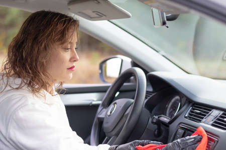 A beautiful young woman driver in a white jacket and black gloves wipes the dust in the car interior with a red rag on a bright warm autumn day. Selective focus. Portraitの写真素材
