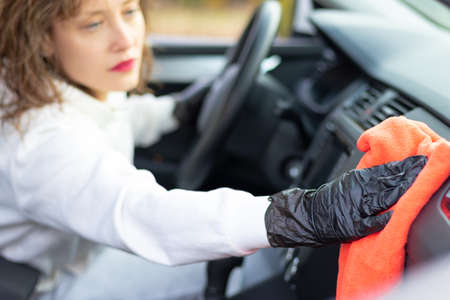 A beautiful young woman driver in a white jacket and black gloves wipes the dust in the car interior with a red rag on a bright warm autumn day. Selective focus. Portraitの写真素材