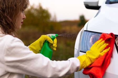 A pretty young woman in a white hoodie and yellow rubber gloves wipes the headlights of a car with a bright red rag on a warm autumn day. Selective focus. Close-upの写真素材