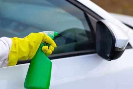 A hand in a yellow rubber glove sprays water from a spray bottle onto a car window on a warm autumn day. Wet cleaning. Selective focus. Close-upの写真素材