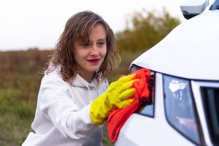 A pretty young woman in a white hoodie and yellow rubber gloves wipes the headlights of a car with a bright red rag on a warm autumn day. Selective focus. Close-upの写真素材