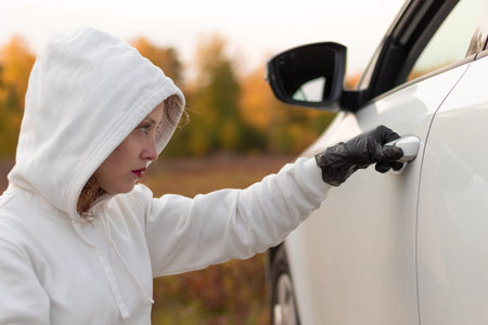 A beautiful young woman in a white sweater with a hood and black gloves opens the car door to steal it. Selective focus. Close-up. Portraitの写真素材