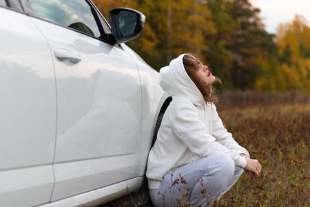 A pretty young woman in a white hoodie and sweatpants sits near a white car with reflection on a warm autumn day against a background of yellow foliage. Selective focus. Portraitの写真素材