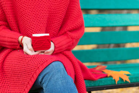 Female hands in a bright red sweater and bracelet hold a red mug with a hot drink on the background of a bench with autumn yellow leaves. Selective focus. Close-upの写真素材