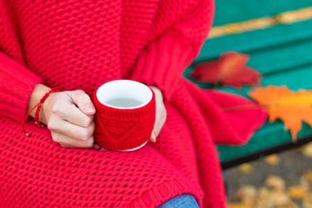 Female hands in a bright red sweater and bracelet hold a red mug with a hot drink on the background of a bench with autumn yellow leaves. Selective focus. Close-upの写真素材