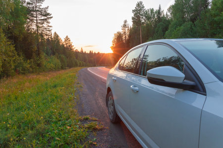 On the side of the road, a part of a white car against the backdrop of a sunset in the forest. Green...