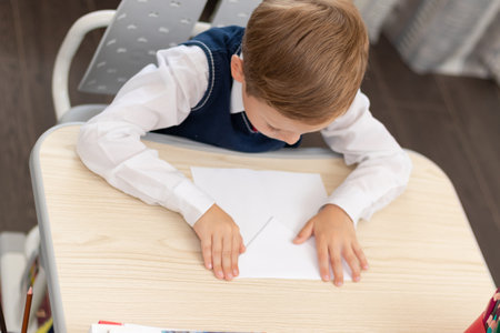 A cute first-grader boy in a school uniform at home while isolated at his desk makes a paper airplane during recess. Selective focus. Portraitの写真素材