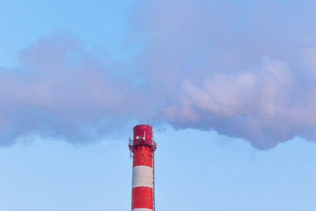 red and white chimney of a factory, from which gray smoke comes out against a blue sky on a sunny...
