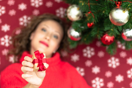 A beautiful young woman in a red sweater holds a box with a gift tied with a red ribbon at home near...