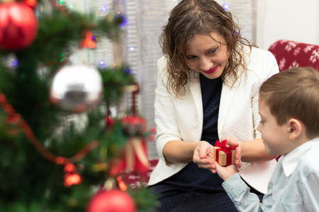 A seven-year-old boy in a smart shirt gives his mother a gift in a box for the New Year against the backdrop of a Christmas tree at home. Selective focus. Portraitの写真素材
