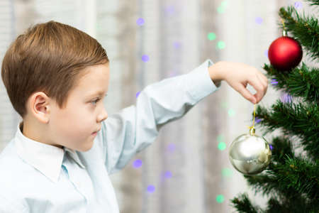 A seven-year-old boy in a blue shirt decorates a Christmas tree with toys on New Year's Eve at home against a background of garlands. Selective focus. Portraitの写真素材