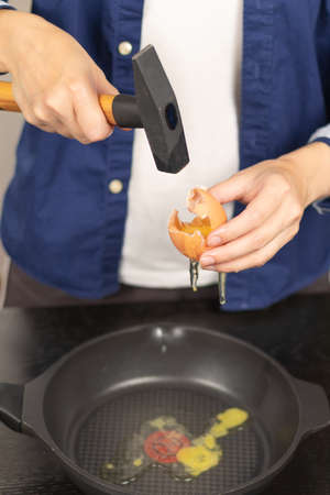 Female hands in a blue shirt break an egg with a hammer at home in the kitchen. Selective focus. Conceptual photo. Close-upの写真素材