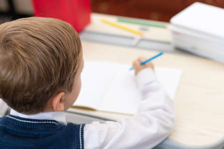 A cute boy first grader in a school uniform does homework while sitting at a desk with a pencil in his hands at home. Remote education. Selective focus. Close-up. Portraitの写真素材