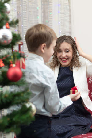 A seven-year-old boy in a smart shirt gives his mother a gift in a box for the New Year against the backdrop of a Christmas tree at home. Selective focus. Portraitの写真素材