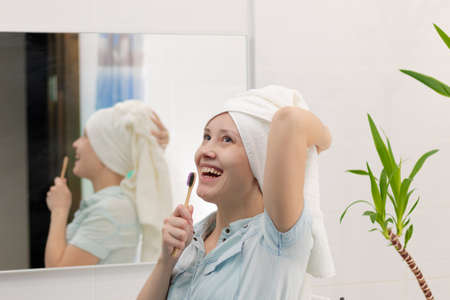 A young pretty woman in a blue bathrobe with a towel on her head in a bright bathroom with a toothbrush in her hand sings songs. Reflection in the mirror with a green houseplant. selective focus. close-upの写真素材