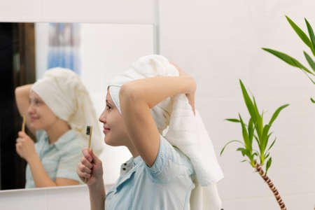 A young pretty woman in a blue bathrobe with a towel on her head in a bright bathroom with a toothbrush in her hand sings songs. Reflection in the mirror with a green houseplant. selective focus. close-upの写真素材