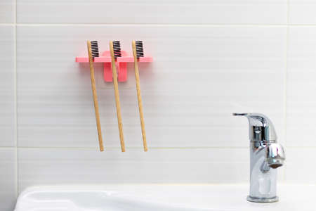 Three natural bamboo toothbrushes with black bristles in a holder against a tile background on the bathroom wall above the washbasin. selective focus. close-upの写真素材