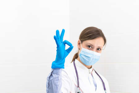 Beautiful young doctor woman in a medical mask and gloves with a stethoscope in the hospital shows hand gestures. selective focus. portrait. close-upの写真素材