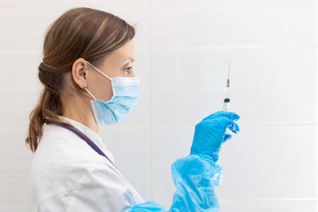 Young woman doctor in a medical mask and gloves with a syringe in her hand and a stethoscope in a hospital during a coronavirus pandemic. selective focus. Portraitの写真素材