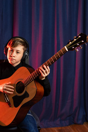 A thirteen-year-old teenager in a black shirt and headphones with a guitar in a music studio records...