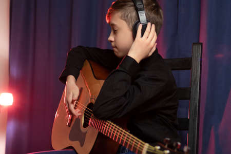 A thirteen-year-old teenager in a black shirt and headphones with a guitar in a music studio records a melody on a purple background with red illumination. selective focus. Portraitの写真素材