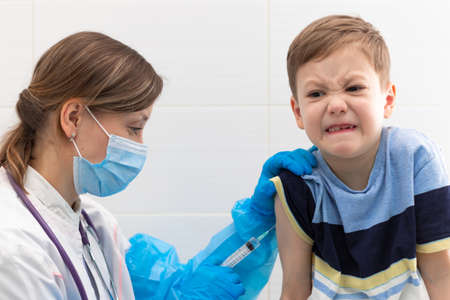 A young female doctor in a medical gown and mask inoculates a boy with a syringe against coronavirus in a hospital during a pandemic. selective focus. Portraitの写真素材