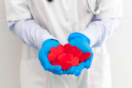 The hands of a doctor in a white medical coat and rubber gloves with a phonendoscope hold a lot of red hearts in the hospital. selective focus. close-upの写真素材