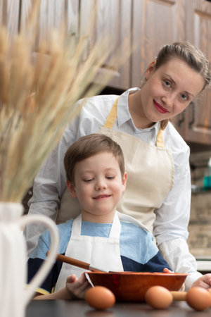 A young beautiful mother with her son in aprons at home in the kitchen prepare dough for baking...