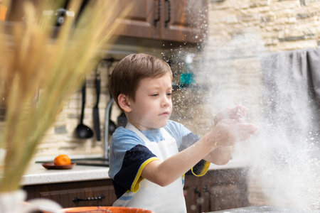 A cute boy of 7 years old in an apron plays with flour at home in the kitchen against the background of kitchen utensils. selective focus. Portraitの写真素材
