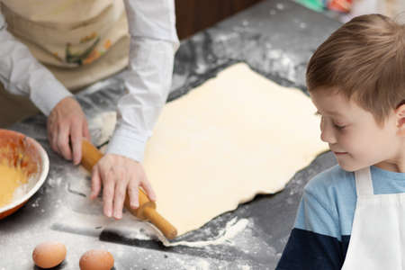 Mom and son in aprons roll out puff pastry for baking on a black kitchen table sprinkled with flour at home with a wooden rolling pin. selective focus. close-upの写真素材