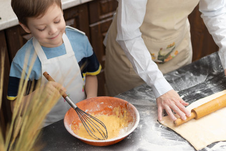Mom and son in aprons roll out puff pastry for baking on a black kitchen table sprinkled with flour at home with a wooden rolling pin. selective focus. close-upの写真素材