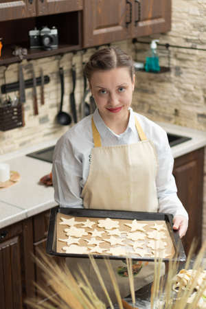 A young pretty woman cook in an apron holds a baking sheet with puff pastry in her hands at home in the kitchen against the background of household utensils. selective focus. Portraitの写真素材