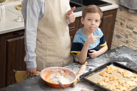 Mom and son try sweet puff pastry lying on parchment on a baking sheet at home in the kitchen. Nearby lies a rolling pin and a bowl. selective focus. portrait. close-upの写真素材