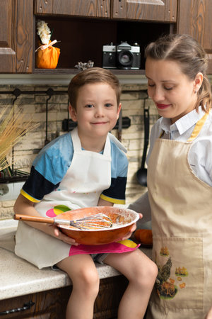 Beautiful young mom cook in an apron with her son prepares cookie dough in a wooden bowl at home in the kitchen. selective focus. portrait. close-upの写真素材
