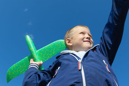 A six-year-old preschooler boy in a blue jacket launches a toy plane in nature against the...