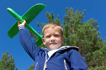 A six-year-old preschooler boy in a blue jacket launches a toy plane in nature against the background of a clear blue sky on a summer day. The bright sun is shining. Portraitの写真素材