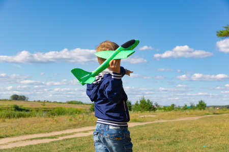A six-year-old preschooler boy in a blue jacket launches a toy plane in a field against a blue sky with clouds on a summer day. The bright sun is shining. sceneryの写真素材