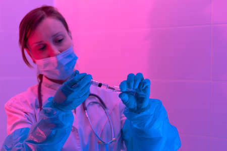 Beautiful young woman doctor with a stethoscope in a protective mask and rubber gloves in a bio laboratory with a syringe takes a sample of the virus. close-up. Pandemic. Portraitの写真素材