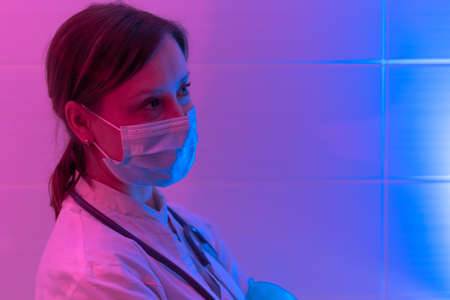 Beautiful young woman doctor with a stethoscope in a protective mask in a bio laboratory on a red-blue background. close-up. Pandemic. Portraitの写真素材