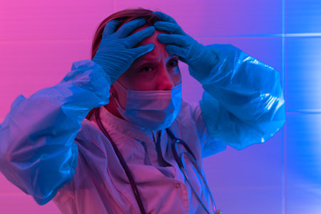 Beautiful young tired woman doctor with a stethoscope in a protective mask and rubber gloves in a bio laboratory on a red-blue background. close-up. Pandemic. Portraitの写真素材