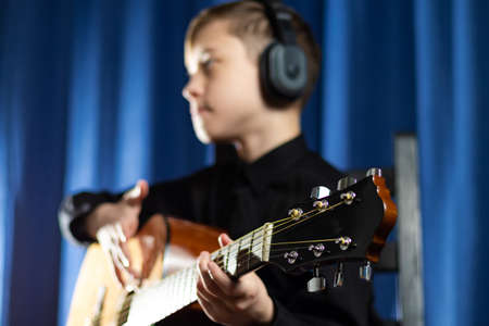 A thirteen-year-old teenager in a black shirt and headphones with a guitar in a music studio records a melody on a blue background. selective focus. Portraitの写真素材
