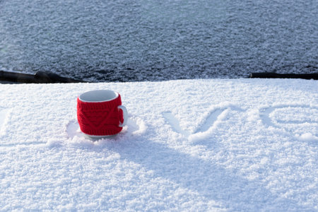 A red mug with a hot drink on a snowy car hood on a bright winter sunny day. The inscription on the...