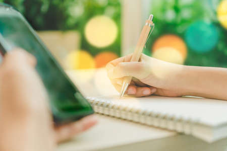 Woman hand using smartphone and writing in notebook  to work or study with cafe shop colorful beautiful bokeh background. Business, financial, trade stock maket and social network concept.の写真素材