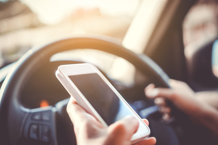 Hand of woman on steering wheel drive a car while using smartphone sunlight background.