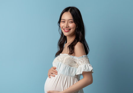 Young pregnant woman in a white dress touching her belly isolated on a blue background.の素材