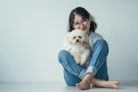 Young Asian woman wearing glasses and jeans is sitting on the floor holding her Poodle dog in front of a white background.の素材